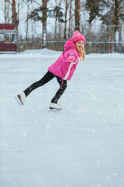 Kleines lächelndes Mädchen, das auf Eis in der rosafarbenen Abnutzung eisläuft. Winter