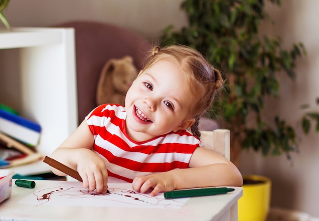 Kleines lächelndes Mädchen sitzt am Tisch und zeichnet mit Filzstiften