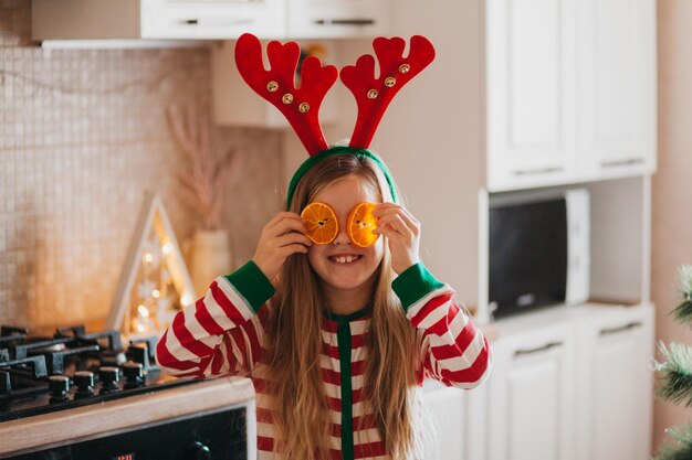 Kleines lächelndes süßes blondes Mädchen in Pyjamas und Geweih hält Fruchthälften in der Nähe ihrer Augen. Kind in der Küche neben dem Weihnachtsbaum. Weihnachten