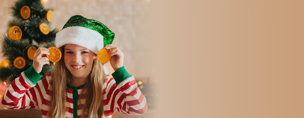 Kleines lächelndes süßes blondes Mädchen in Schlafanzug und Geweih hält Fruchthälften in der Nähe ihrer Augen Kind in der Küche neben dem Weihnachtsbaum Weihnachten