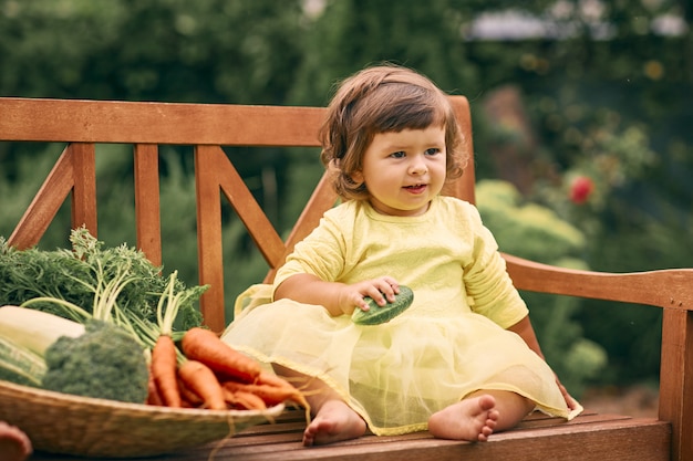 Kleines lockiges Mädchen in einem gelben Kleid, in einem grünen Garten, hält eine Gurke in der Hand, sitzt auf einer Bank neben einem großen Korb mit Gemüse. Gesundes Essen, grünes vegetarisches Essen.