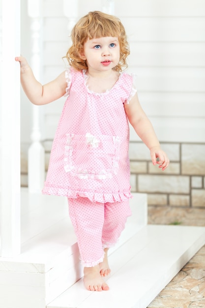 Kleines lockiges Mädchen in einem rosa Kleid und bloßen Füßen, die aus dem Haus kommen und die Treppe hinuntergehen