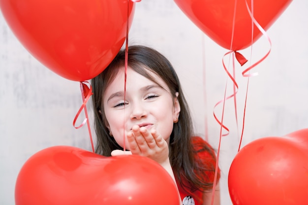 Kleines lustiges freches mädchengesicht, das einen kuss hautnah zwischen roten herzförmigen luftballons gibt
