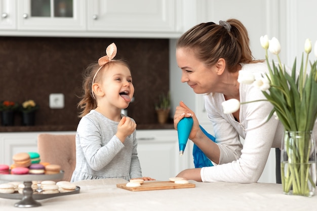 Kleines lustiges Kindermädchen und ihre Mutter backen Macarons und Kekse in der weißen Küche baking