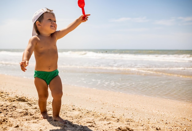 Kleines lustiges lustiges Kind, sammelt Muscheln und Kieselsteine im ruhigen blauen Meer auf einem sandigen Boden unter der heißen Sommersonne in einem hellen Urlaub