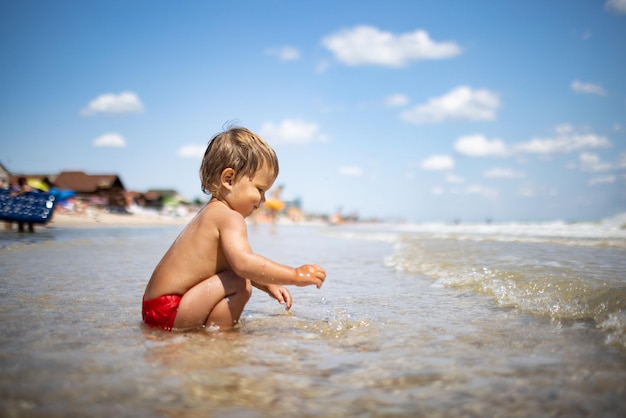 Kleines lustiges lustiges Kind sammelt Muscheln und Kieselsteine im ruhigen blauen Meer auf einem sandigen Boden unter der heißen Sommersonne in einem hellen Urlaub