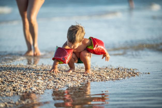Kleines lustiges lustiges Kind, sammelt Muscheln und Kieselsteine im ruhigen blauen Meer auf einem sandigen Boden zusammen mit einer fürsorglichen, freundlichen, schönen Mutter unter heißer Sommersonne in einem hellen Urlaub