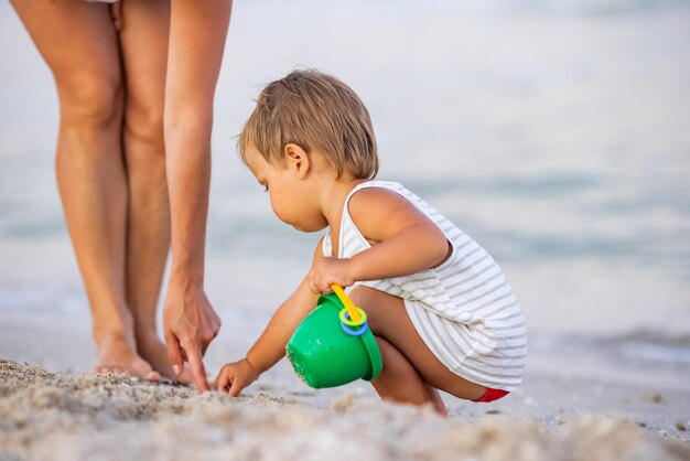 Kleines lustiges lustiges Kind, sammelt Muscheln und Kieselsteine im ruhigen blauen Meer auf einem sandigen Boden zusammen mit einer fürsorglichen, schönen Mutter unter heißer Sommersonne in einem hellen Urlaub