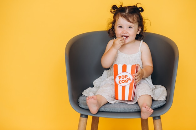 Kleines lustiges Mädchen, das zu Hause auf einem Stuhl isst Popcorn lächelt und sitzt und einen Film aufpasst