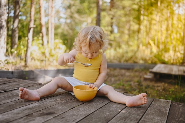 Kleines lustiges süßes blondes Mädchenkindkleinkind mit schmutziger Kleidung und Gesicht, das Babynahrungsfrucht isst oder