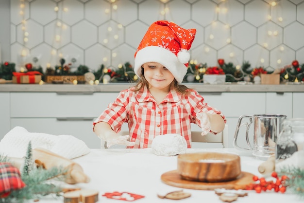 Kleines Mädchen 3 Jahre alt in roter Weihnachtsmütze und kariertem Hemd bereitet Teig für Lebkuchen in der Küche vor