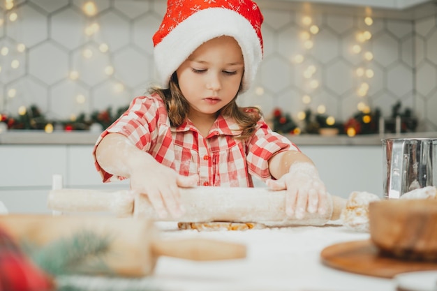 Kleines Mädchen 3 Jahre alt in roter Weihnachtsmütze und kariertem Hemd rollen Teig für Lebkuchen in der Küche aus