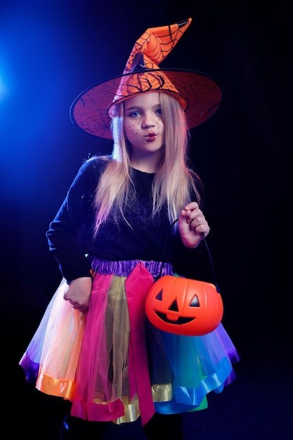 Kleines Mädchen als Hexe mit Kürbis auf einem dunklen Hintergrund im Licht mehrfarbiger Scheinwerfer gekleidet