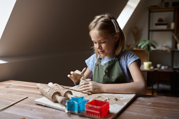 Kleines Mädchen arbeitet mit Ton am Tisch, Kind in der Werkstatt. Unterricht an der Kunstschule. Junger Meister des Volkshandwerks, angenehmes Hobby, glückliche Kindheit