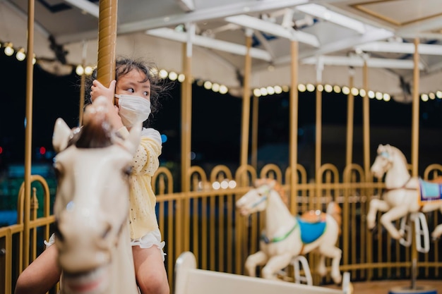 Kleines Mädchen auf Karussellpferd mit gesunder Gesichtsmaske zum Schutz vor Viren und Umweltverschmutzung.