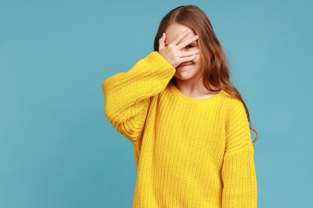 Kleines Mädchen bedeckt das Gesicht mit der Hand, die durch die Finger schaut, neugierig und schüchtern, das Geheimnis zu beobachten, das einen gelben, lässigen Pullover trägt Studioaufnahme isoliert auf blauem Hintergrund