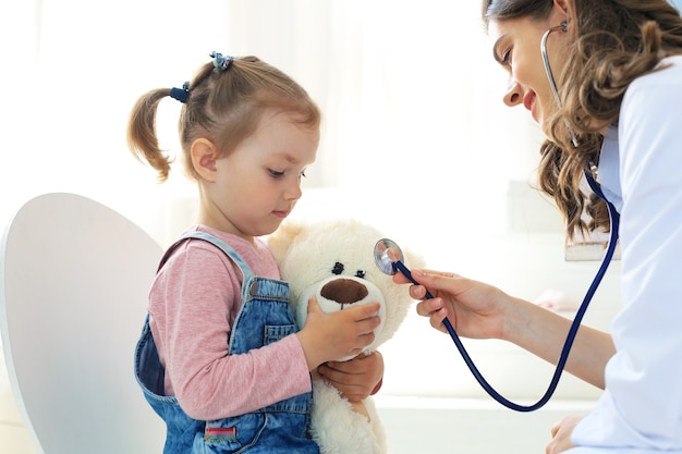 Kleines Mädchen beim Arzt zur Untersuchung. Doktor, der spielerisch den Herzschlag eines Teddybären überprüft.