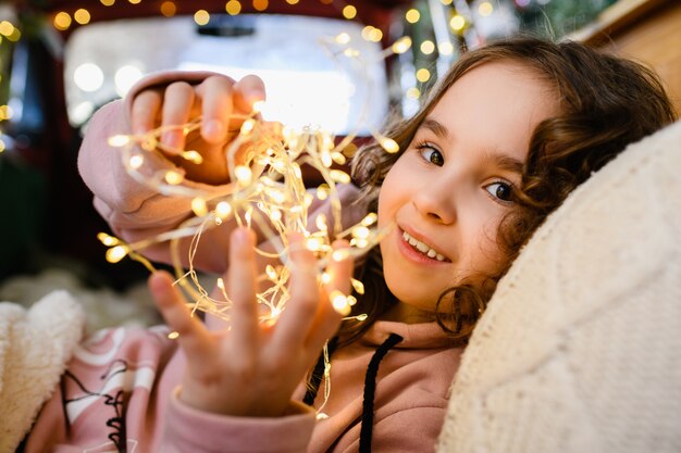 Kleines Mädchen beleuchtetes Gesicht, das warme gelbe Lichter der Girlande am Weihnachts- und Neujahrshintergrund hält