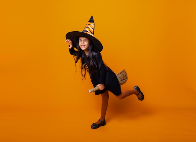 Kleines Mädchen, das als Halloween-Hexe in einem schwarzen Kleid und Hut verkleidet ist, fliegt auf einem Besenstiel auf gelbem Hintergrund.