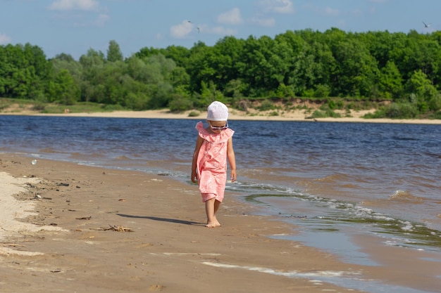 Kleines Mädchen, das am Sommerstrand spaziert, der von Wellen umspült wird, mit schöner Landschaft im Hintergrund