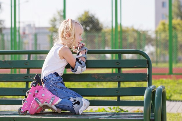 Kleines Mädchen, das auf Bank im Spielplatz in Rollschuhen sitzt