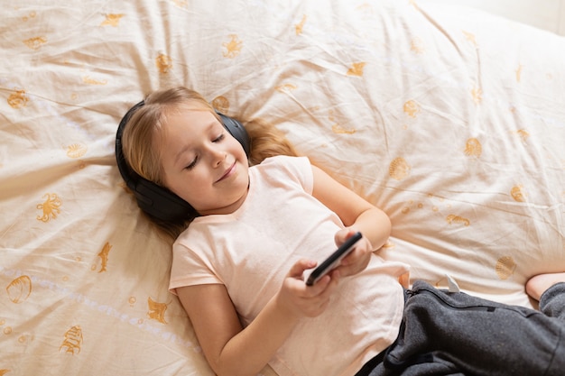 Kleines Mädchen, das auf dem Bett mit kabellosen Kopfhörern und Telefon liegt