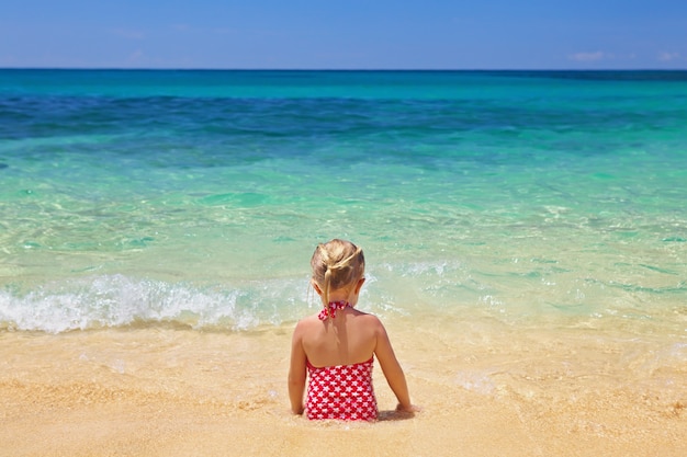 Foto kleines mädchen, das auf dem sandstrand sitzt, schaut auf den blauen ozean