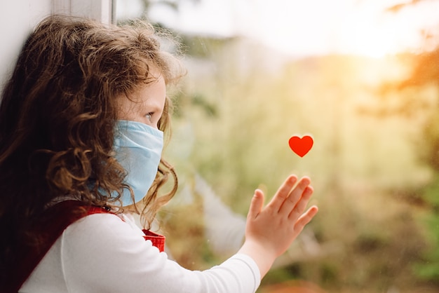 Kleines Mädchen, das blaue medizinische Gesichtsmaske trägt, die auf der Schwelle mit dem kleinen roten Herzen am Fenster sitzt, um Ärzten und Krankenschwestern Dankbarkeit für Hilfe im Kampf gegen die Krankheit zu zeigen. Covid-19