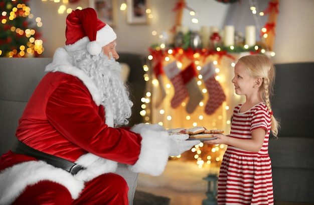 Kleines Mädchen, das den Weihnachtsmann mit leckeren Keksen im Zimmer mit schönen Weihnachtsdekorationen behandelt