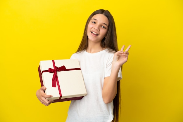 Kleines Mädchen, das ein Geschenk über isoliertem gelbem Hintergrund hält und lächelt und Victory-Zeichen zeigt