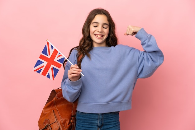 Kleines Mädchen, das eine britische Flagge lokalisiert auf rosa Hintergrund hält, der starke Geste tut