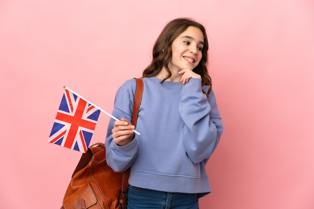 Kleines Mädchen, das eine britische Flagge lokalisiert auf rosa Hintergrund hält, der zur Seite schaut und lächelt