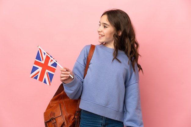 Kleines Mädchen, das eine britische Flagge lokalisiert auf rosa Hintergrund hält, der zur Seite schaut und lächelt