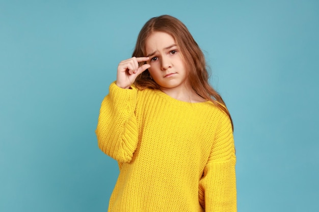 Kleines Mädchen, das eine kleine Geste macht, mit unzufriedenem, beschwörendem Ausdruck aussieht, zeigt ein Minimum und trägt einen gelben Pullover im Freizeitstil. Studioaufnahme im Innenbereich isoliert auf blauem Hintergrund.