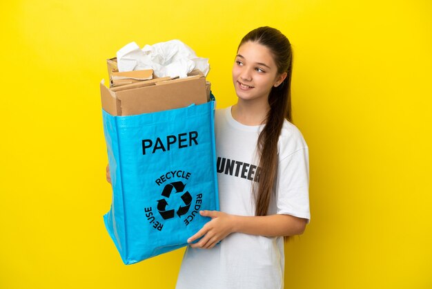 Kleines Mädchen, das eine Recyclingtüte voller Papier hält, um über isolierten gelben Hintergrund zu recyceln, der zur Seite schaut und lächelt