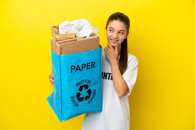 Kleines Mädchen, das eine Recyclingtüte voller Papier hält, um über isolierten gelben Hintergrund zu recyceln, der zur Seite schaut und lächelt