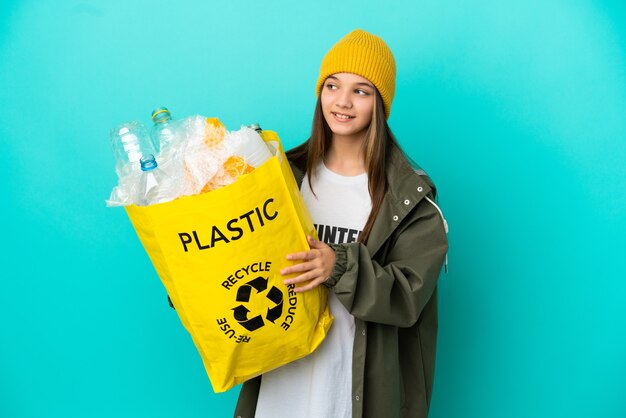 Kleines Mädchen, das eine Tasche voller Plastikflaschen hält, um über eine isolierte blaue Wand zu recyceln, die zur Seite schaut und lächelt