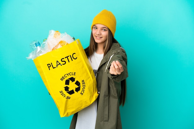 Kleines Mädchen, das eine Tasche voller Plastikflaschen hält, um über isolierte blaue Oberfläche zu recyceln und Geldgeste zu machen