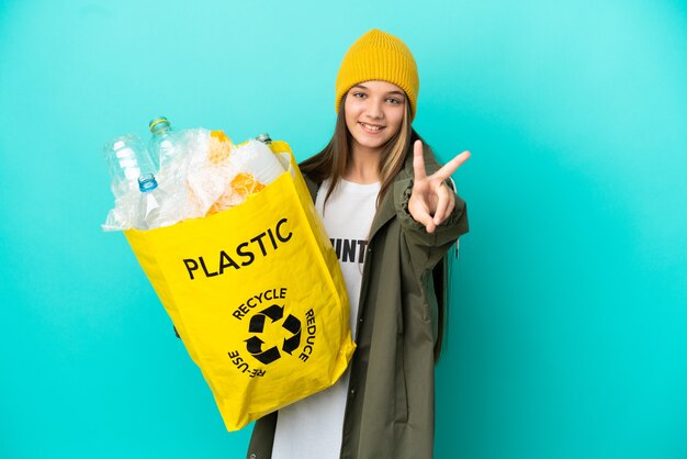 Kleines Mädchen, das eine Tasche voller Plastikflaschen hält, um über isoliertem blauem Hintergrund zu recyceln, lächelt und zeigt Victory-Zeichen