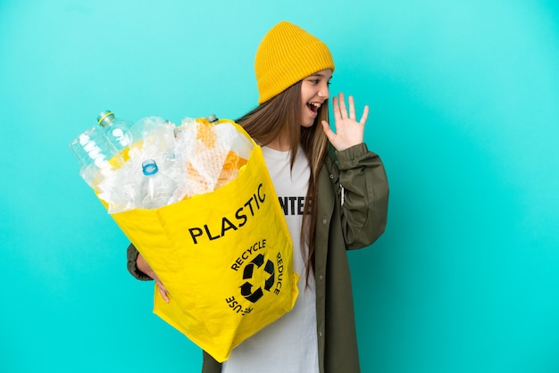 Foto kleines mädchen, das eine tasche voller plastikflaschen hält, um über isoliertem blauem hintergrund zu recyceln, schreit mit weit geöffnetem mund zur seite