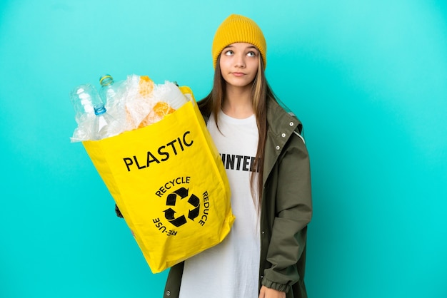 Kleines Mädchen, das eine Tasche voller Plastikflaschen zum Recyceln über isoliertem blauem Hintergrund hält und nach oben schaut