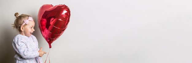 Kleines Mädchen, das einen Herzluftballon hält und es auf einem hellen Hintergrund betrachtet. Konzept für Valentinstag, Geburtstag. Banner.