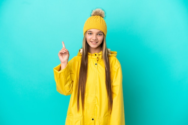 Kleines Mädchen, das einen regendichten Mantel über isoliertem blauem Hintergrund trägt und auf eine großartige Idee hinweist