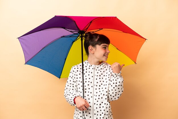 Kleines Mädchen, das einen Regenschirm hält, der auf beige Wand lokalisiert ist, die zur Seite zeigt, um ein Produkt zu präsentieren