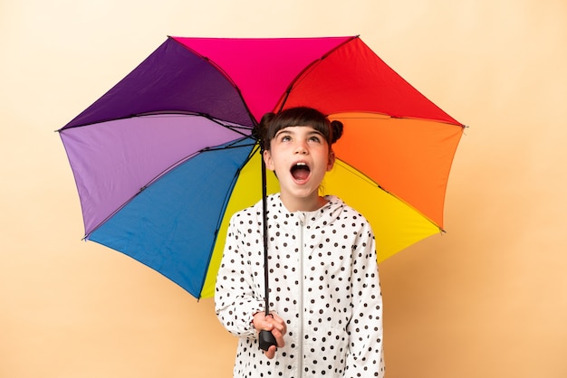 Kleines Mädchen, das einen Regenschirm lokalisiert auf beige Wand hält, die oben und mit überraschtem Ausdruck schaut