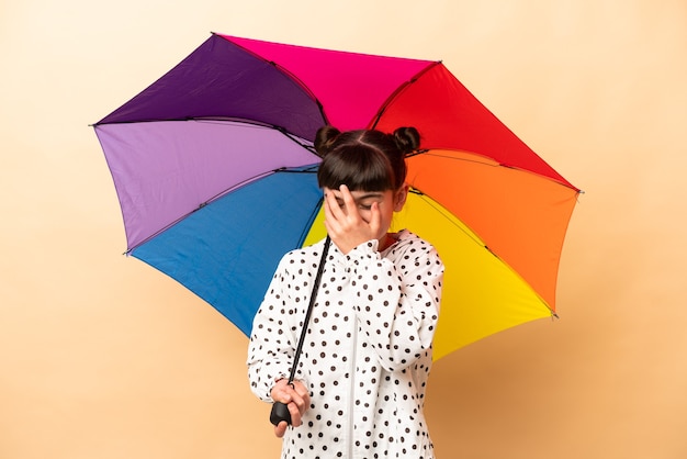 Kleines Mädchen, das einen Regenschirm lokalisiert auf beige Wand lachend hält