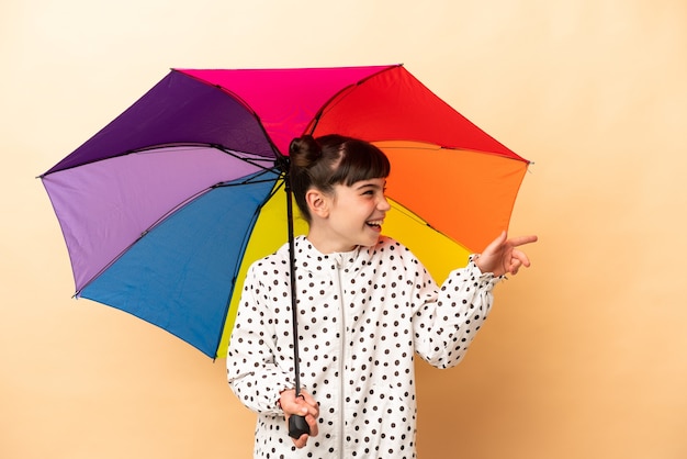 Kleines Mädchen, das einen Regenschirm lokalisiert auf beigem Zeigefinger zur Seite hält und ein Produkt präsentiert