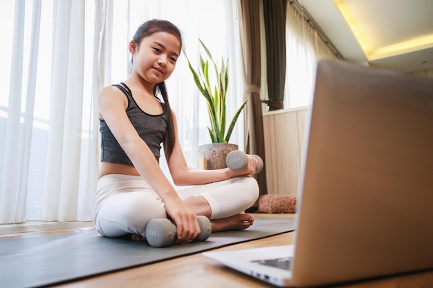 Kleines Mädchen, das Hantelübungen und Online-Lernen mit einem Laptop auf einer Yogamatte macht