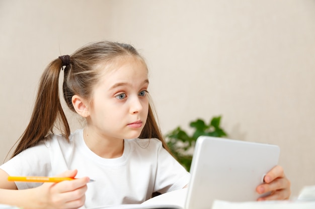 Kleines mädchen, das hausaufgaben zu hause am tisch macht. das kind ist zu hause unterrichtet. ein mädchen mit hellem haar führt eine aufgabe online mit einem laptop und einem tablet-computer aus.