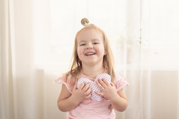 Kleines Mädchen, das Herz hält. Kleines herzförmiges Kissen, das von einem kleinen kaukasischen Mädchen gehalten wird, das unschuldig lächelt, zu Hause. Konzept von Kind, Liebe und Glück, Freundlichkeit, Geschenk, handgemachter Valentinsgruß, Familie